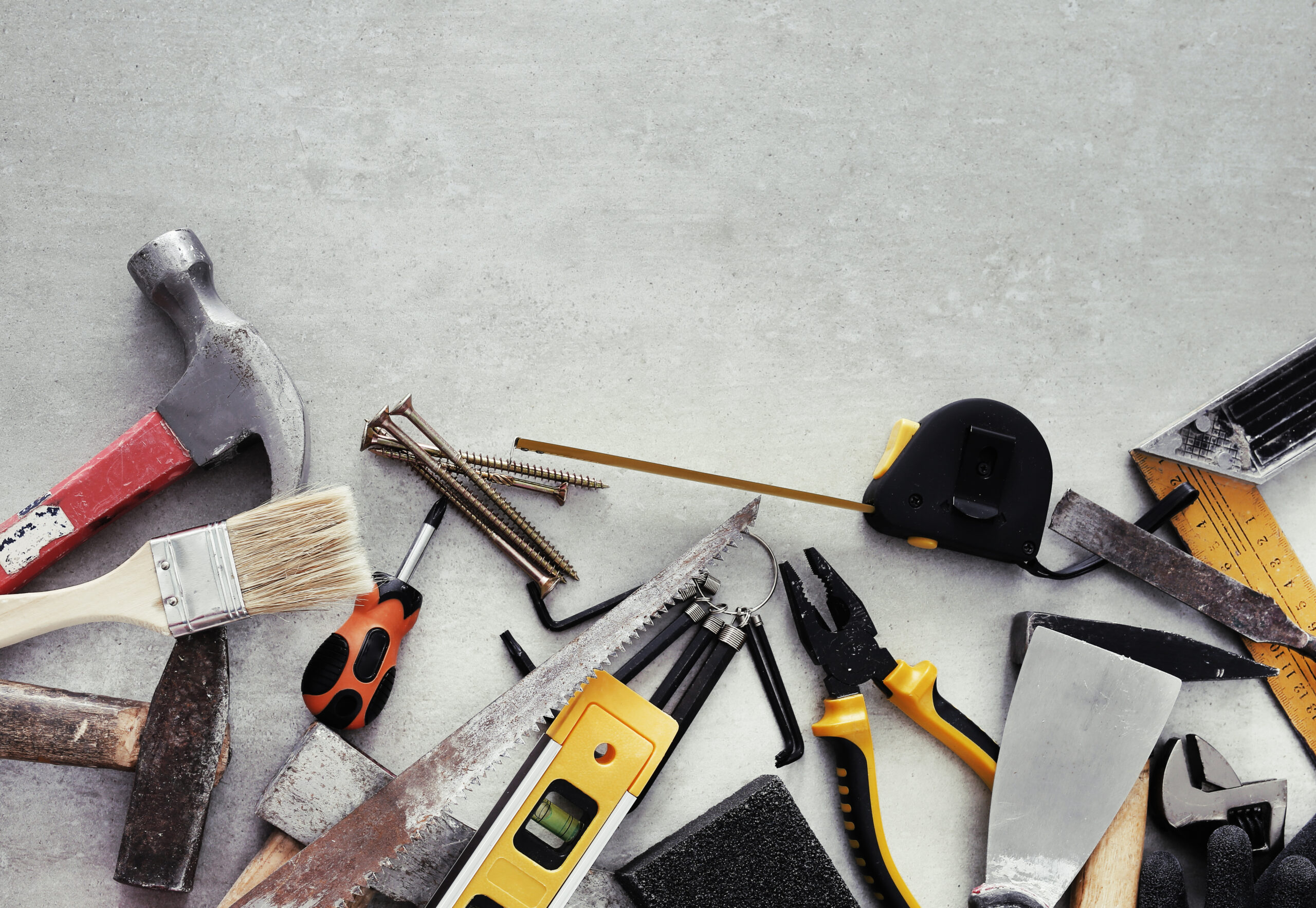 Das Bild zeigt mehrere Werkzeuge wie Hammer, Pinsel, Schrauben, Schraubenziehe und Zangen, die auf einem grauen Betonboden liegen.